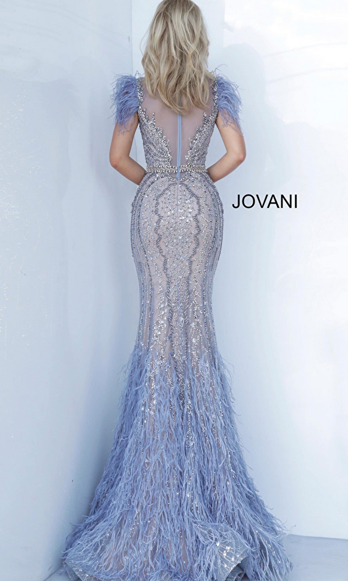 A woman wears the Jovani Light Blue Feather Long Pageant Dress 02326, a fitted floor-length gown with sequins, a mermaid skirt, sheer back, and feathered sleeves as she faces a white wall.